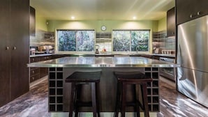 A modern kitchen with a large island, stainless steel appliances, and a view of trees through the windows. - Celadon Holiday House @ PALM COVE (Palm Cove)