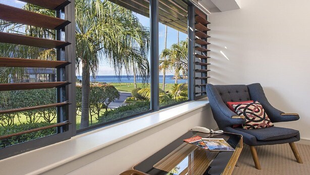 A room with a large window overlooking a landscaped area and the ocean. There is a chair with a cushion and a small table with a magazine and a phone. - Shoreline 501 Sebel Kiama (Kiama)