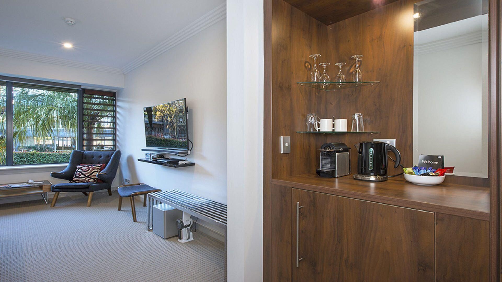 Een moderne hotelkamer met een groot raam, een comfortabele zithoek, een flatscreen televisie en een houten kastje met een koffiezetapparaat en glazen.