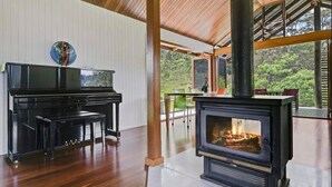 A spacious room with a piano, a fireplace, and large windows offering a view of greenery. - Off grid tranquillity with beautiful views and no other house in sight (Mapleton)