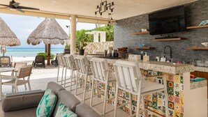Een strandbar met een televisie, rieten parasols en een kleurrijke betegelde muur.