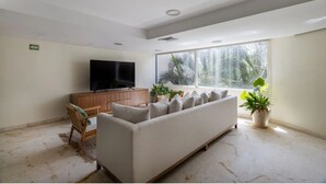 Een woonkamer met een bank, televisie, houten kast en grote ramen met planten.