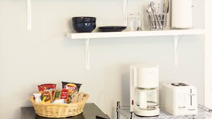 A kitchenette with a black refrigerator, microwave, and coffee maker on a shelf, a basket of snacks, and a painting of palm trees on the wall.