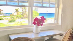 Una habitación con vista al océano, un jarrón blanco con flores en el alféizar de una ventana y dos sillones de mimbre.