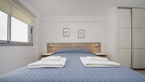 Un lit bien fait, recouvert d’une couette bleue, avec deux serviettes blanches pliées posées sur le lit, une tête de lit en bois et une fenêtre avec des stores.
