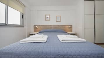 A neatly made bed with a blue quilt, two white towels folded on the bed, a wooden headboard, and a window with blinds.