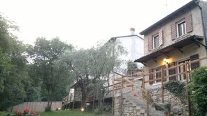A stone building with a wooden staircase leading to a balcony, surrounded by greenery and a gravel driveway.