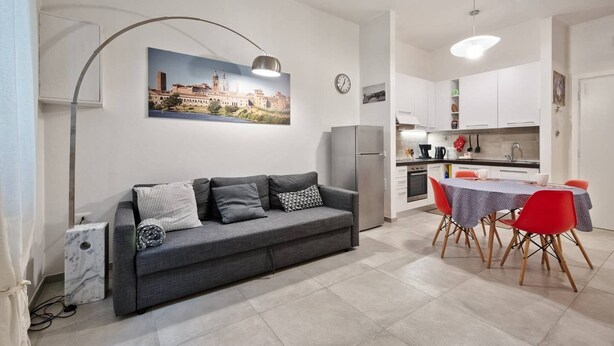 A modern living room with a grey sofa, a dining table with red chairs, a kitchen area, and a cityscape painting on the wall.