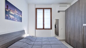 A bedroom with a bed, a window, a wardrobe, and a painting on the wall.
