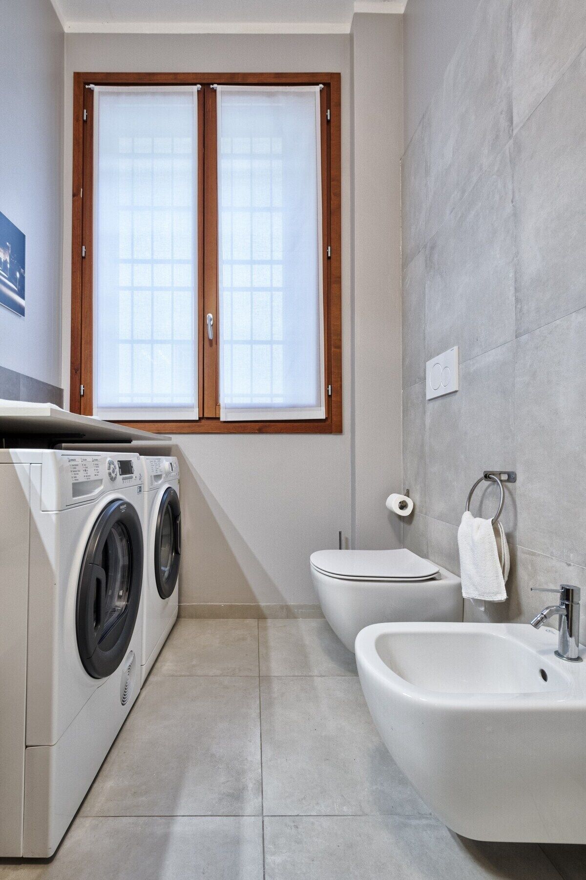A compact bathroom with a washing machine, bidet, and a window with wooden frames.
