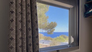 Janela com vista para o mar e árvores, cortinas com padrão geométrico e parede branca.