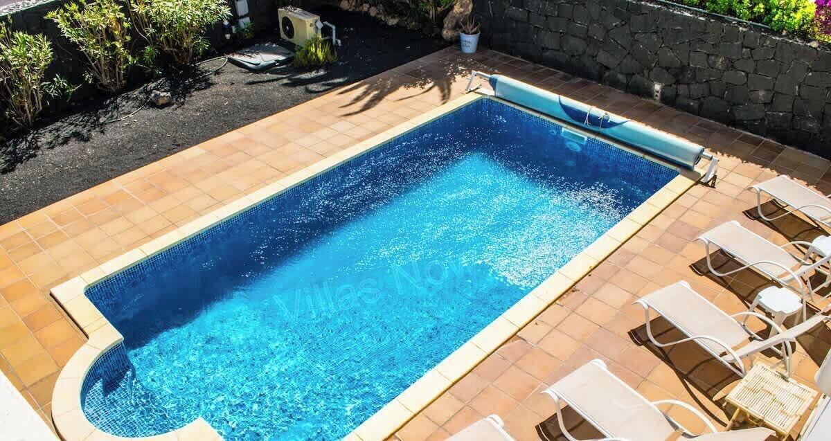 A rectangular swimming pool with a tiled deck, surrounded by a stone wall and potted plants.