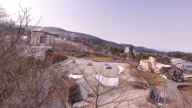A parking lot with several cars, buildings in the background, and a hilly landscape. - Miharashinoyado Kokyo (Yamagata)