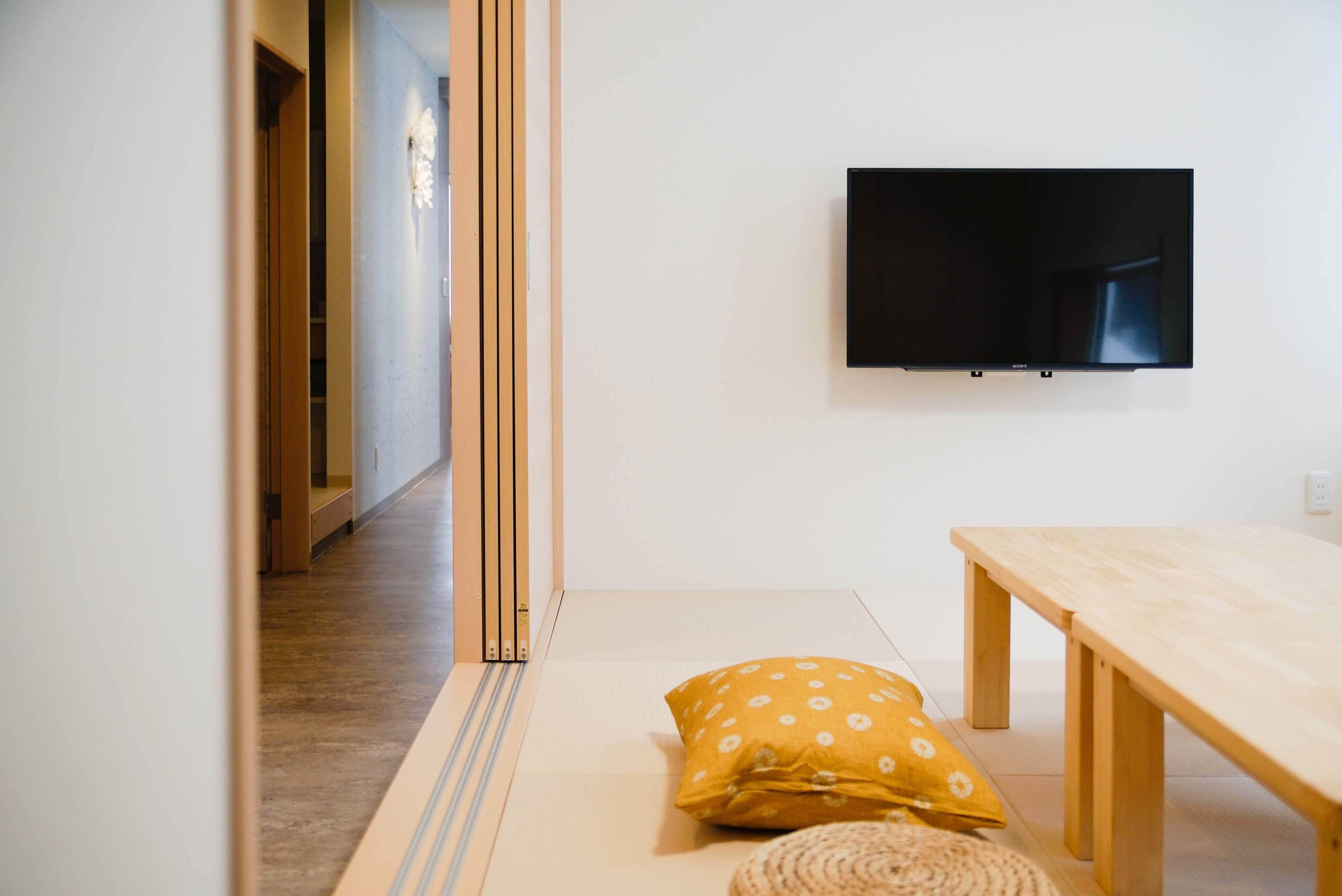 A minimalist room with tatami flooring, a low wooden table, a yellow pillow, and a mounted TV.