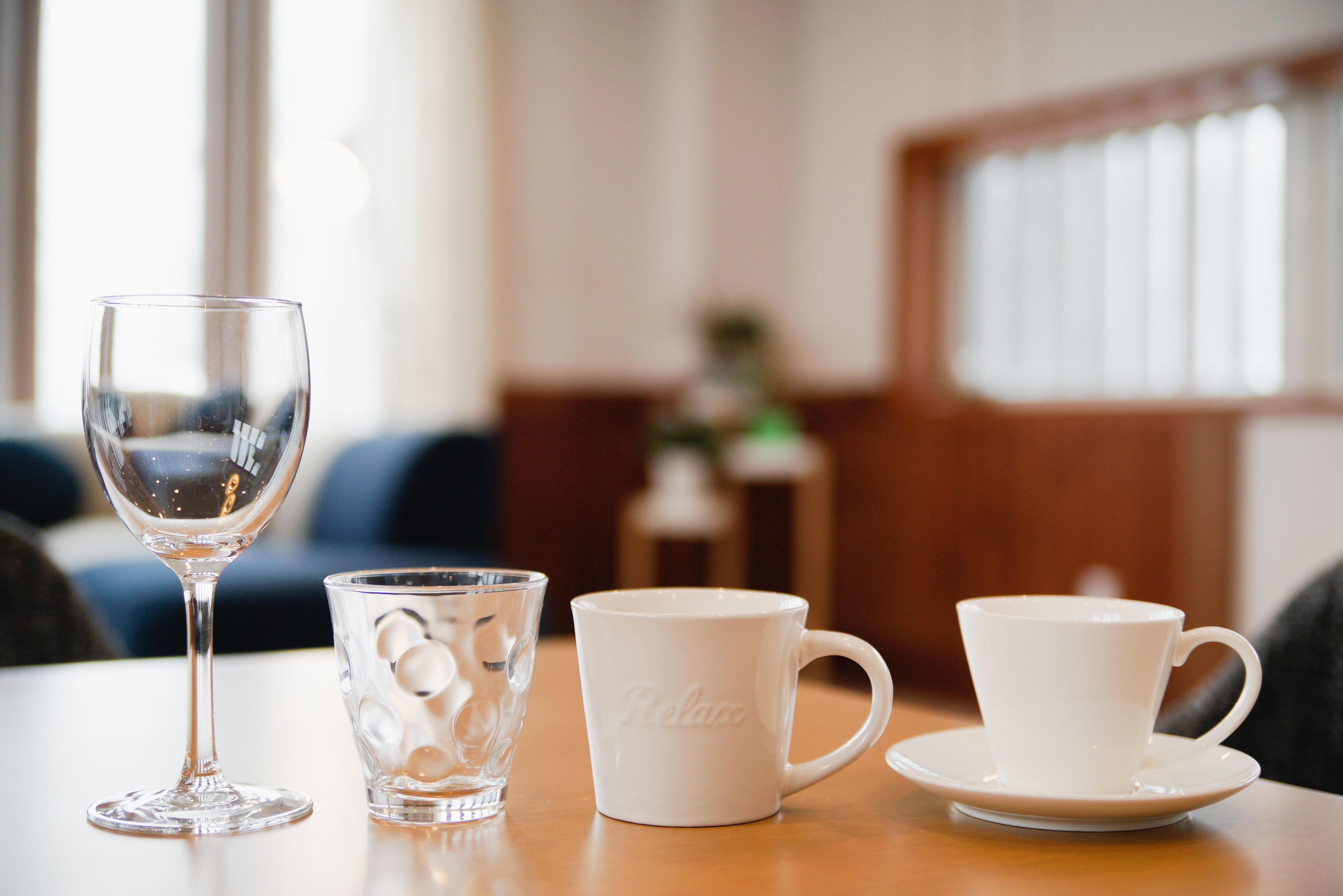 A table with a wine glass, a glass with ice cubes, and two white cups on a wooden table in a room with a sofa and a window in the background.