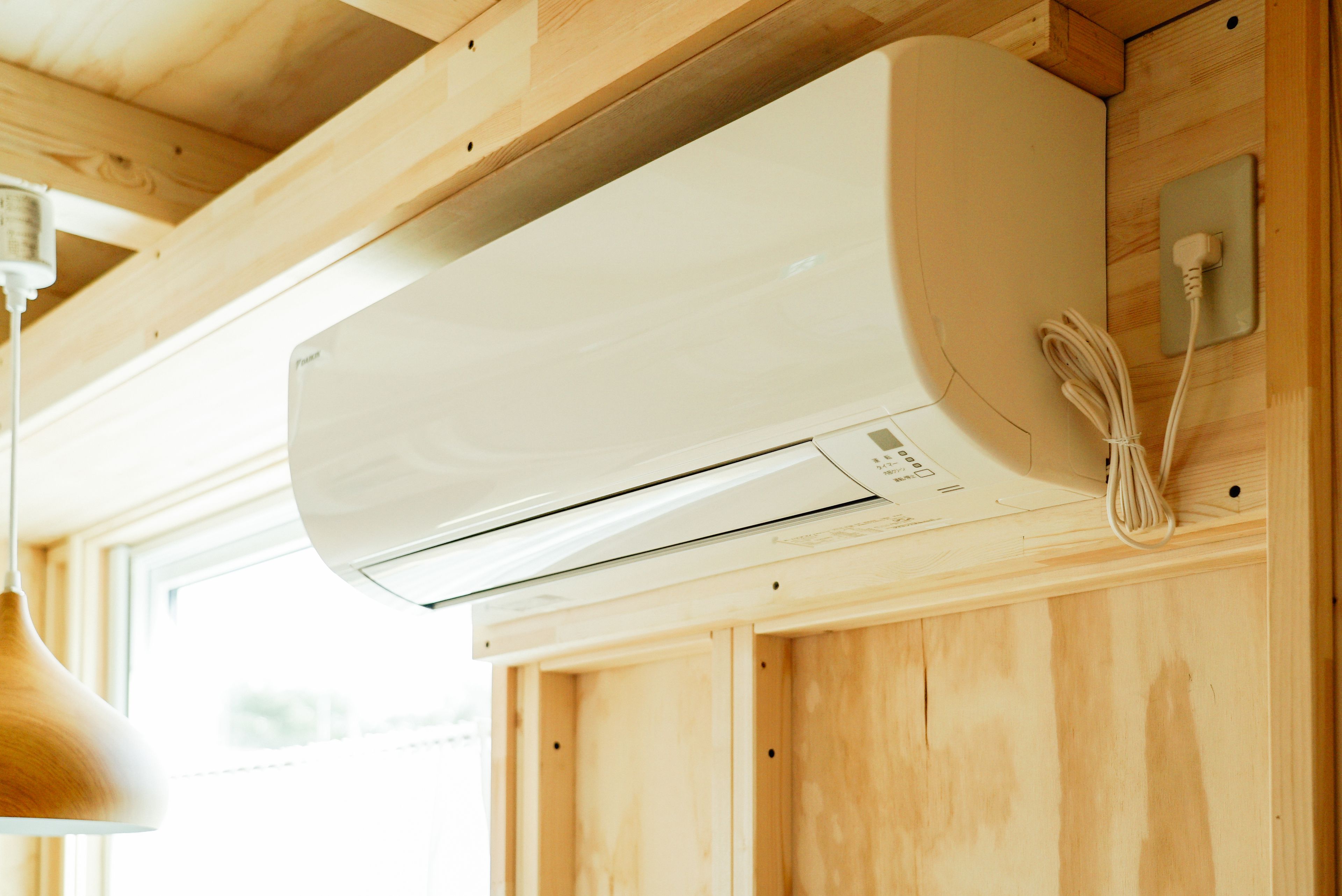 En hvid aircondition-enhed monteret på en trævæg, med strømkabel tilsluttet.