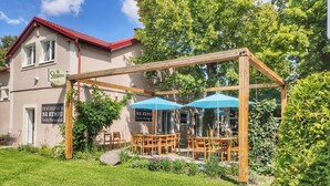A restaurant with outdoor seating under a wooden pergola, surrounded by greenery and a building with a red roof.
