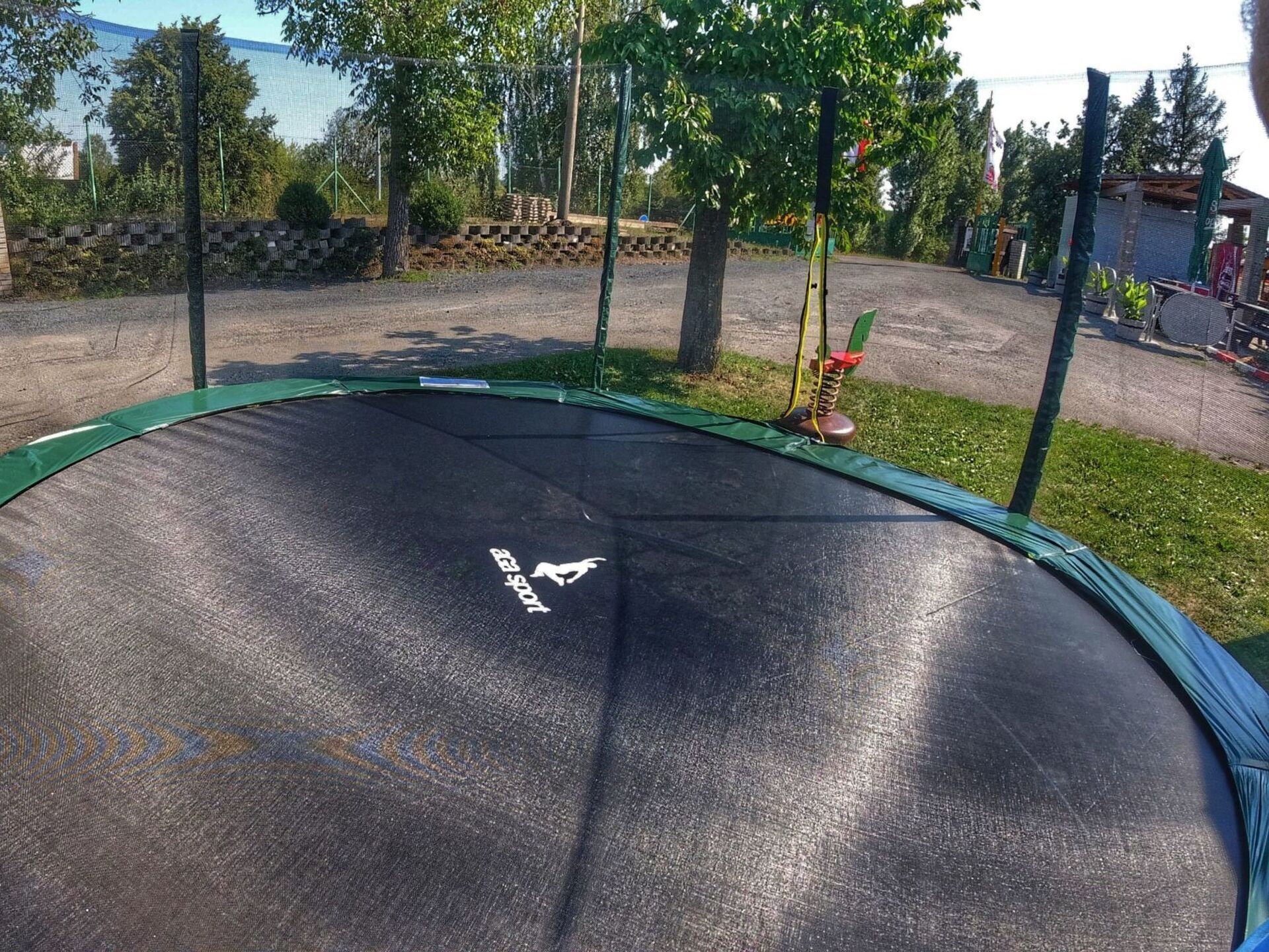 Ein Trampolin mit Sicherheitslogo, umgeben von Bäumen und einem Kiesbereich.