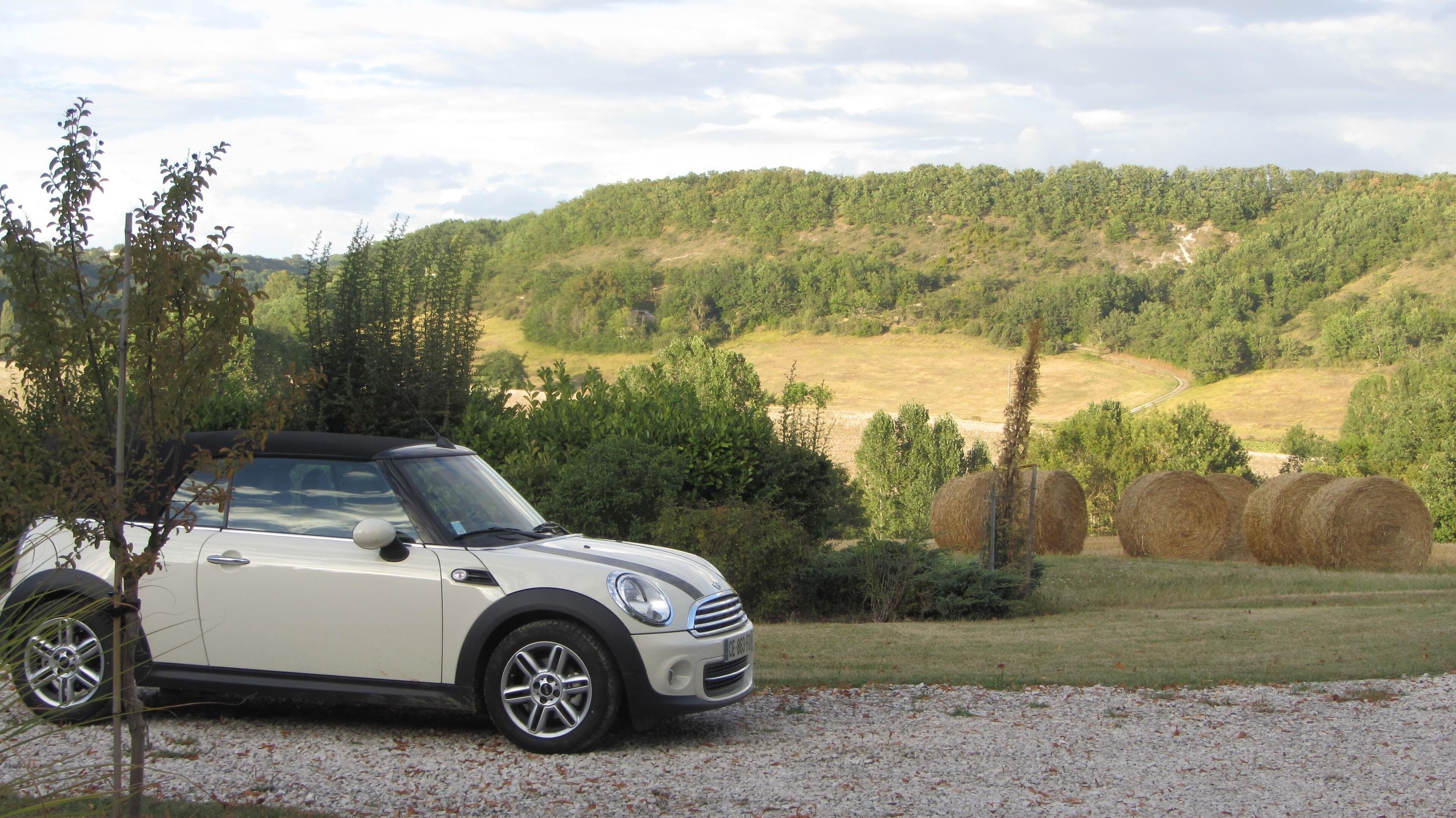 A white convertible car parked on a gravel path with hay bales in the background and a forested hill.