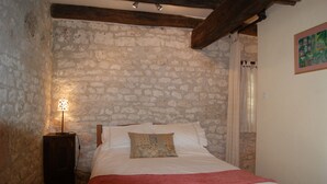 A bedroom with a stone wall, a wooden ceiling beam, a bed with white bedding and a red blanket, and a nightstand with a lamp.