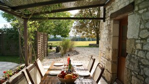 A wooden table set for four with a bowl of fruit, plates, and glasses, under a pergola with a stone wall backdrop.