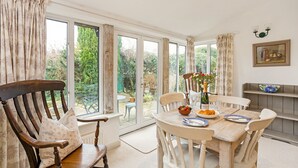 A dining area with a table set for four, surrounded by wooden chairs, a wooden bench, and a view of the garden through large windows. - Cornerstone Cottage -  a cotswold cottage that sleeps 2 guests  in 1 bedroom (Chipping Campden)