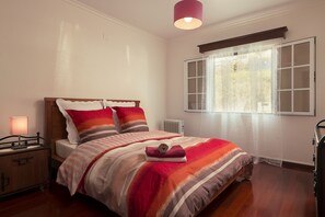 Ein ordentlich bezogenes Bett mit gestreifter Bettwäsche und Kissen, ein Nachttisch mit Lampe und ein Fenster mit durchsichtigen Vorhängen.