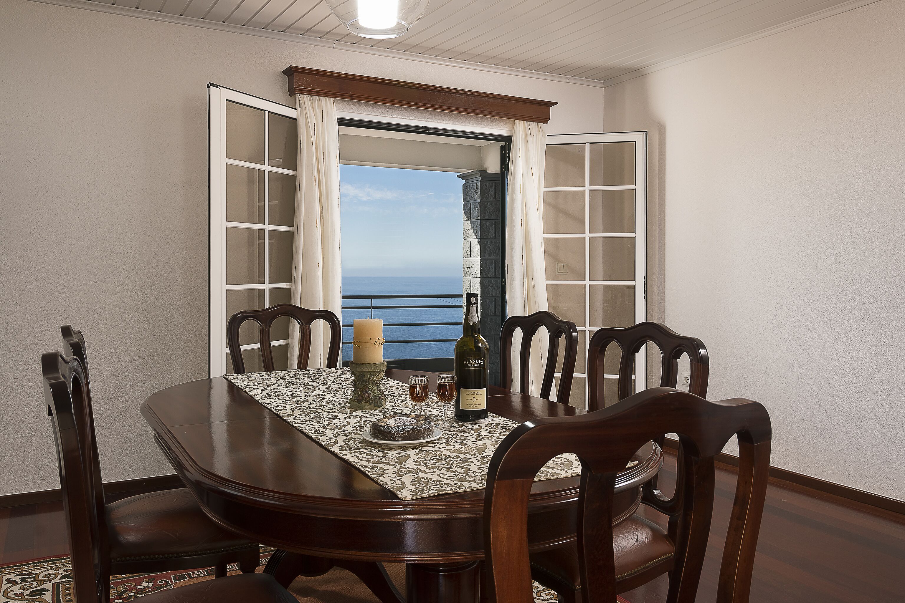 A dining area with a table set for four, a candle, a bottle, and a view of the ocean through an open door.