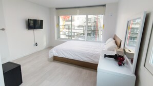 A modern hotel room with a large bed, a flat-screen TV mounted on the wall, a window with a view of buildings, and a small white dresser. - Kpop Haus (Seoul)