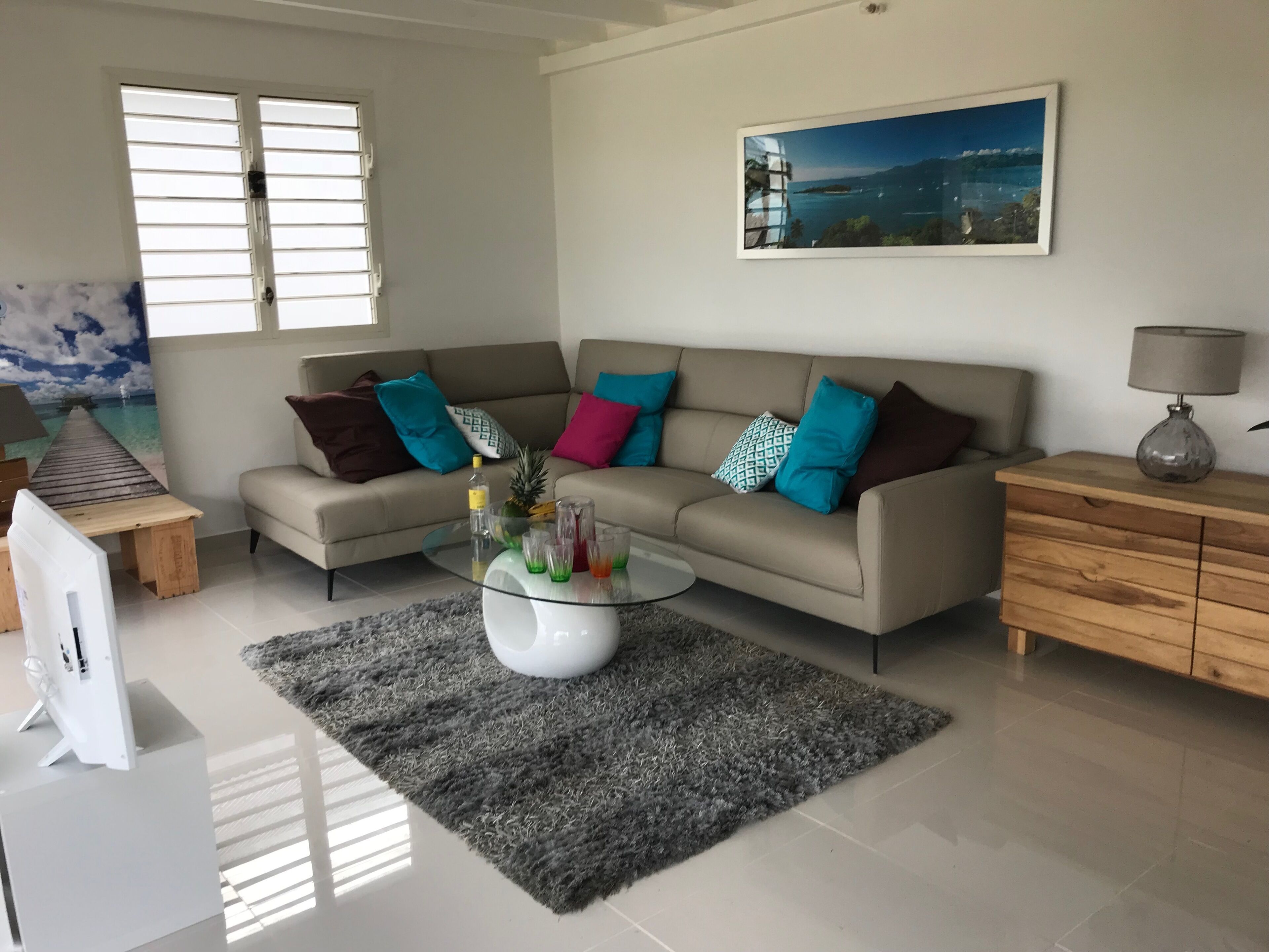 Una sala de estar con un sofá color crema, cojines de colores, una mesa de centro de vidrio y una mesita de noche de madera.