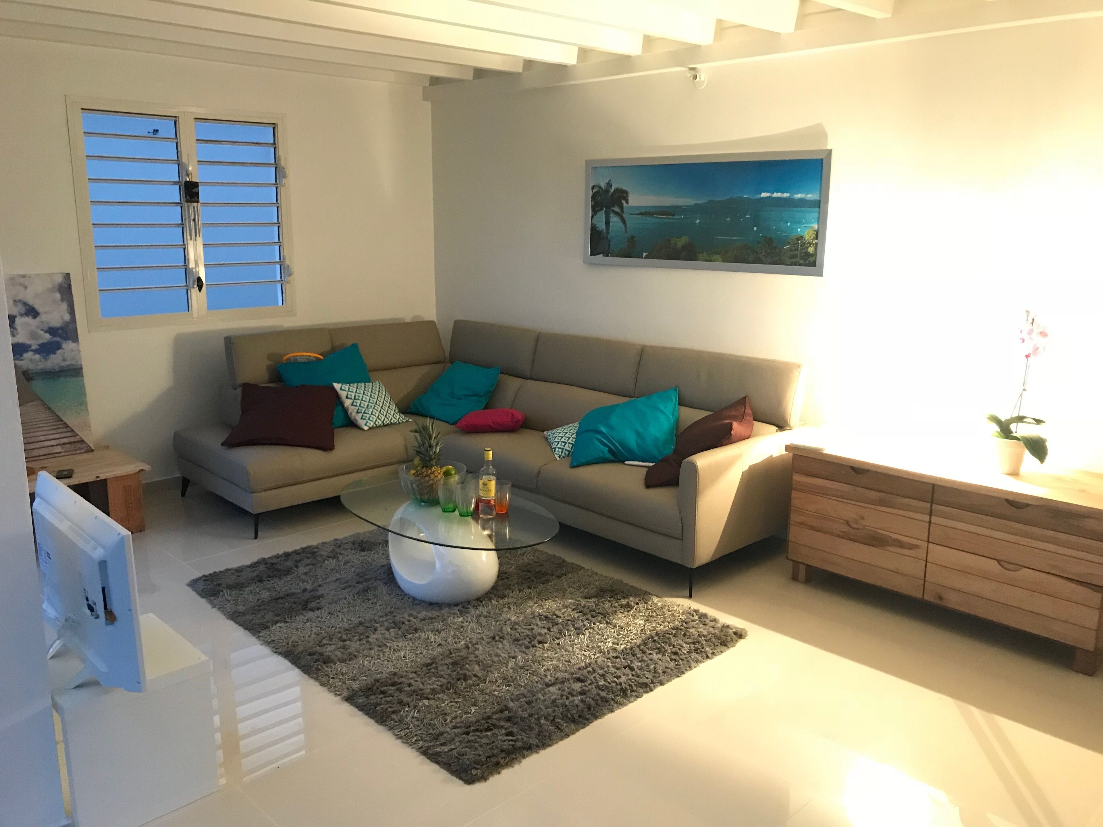 A modern living room with a beige sofa, a glass coffee table, and a view of the ocean through a window.