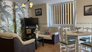 Een woonkamer met een bank, een televisie, een klein tafeltje en een eethoek met stoelen.