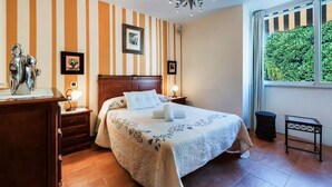 Hotelkamer met een bed, houten hoofdeinde, nachtkastjes, een dressoir, een kroonluchter, een raam met uitzicht op groen en gestreepte wanden.