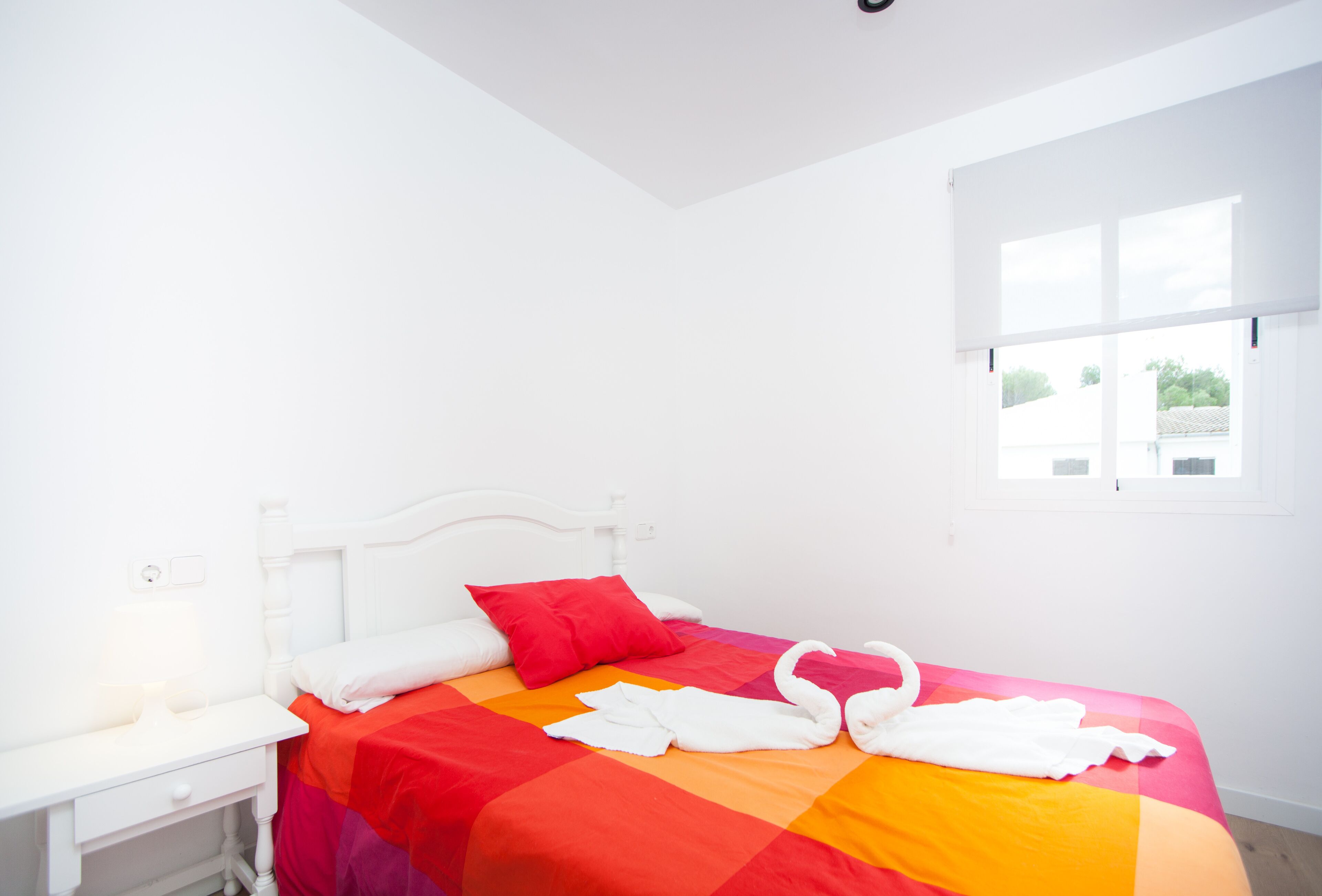A bright, white room with a bed featuring a colorful bedspread and two red pillows.