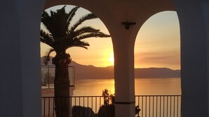 Un balcon offrant une vue sur le coucher de soleil au-dessus d’une étendue d’eau, le tout encadré par des arches architecturales.