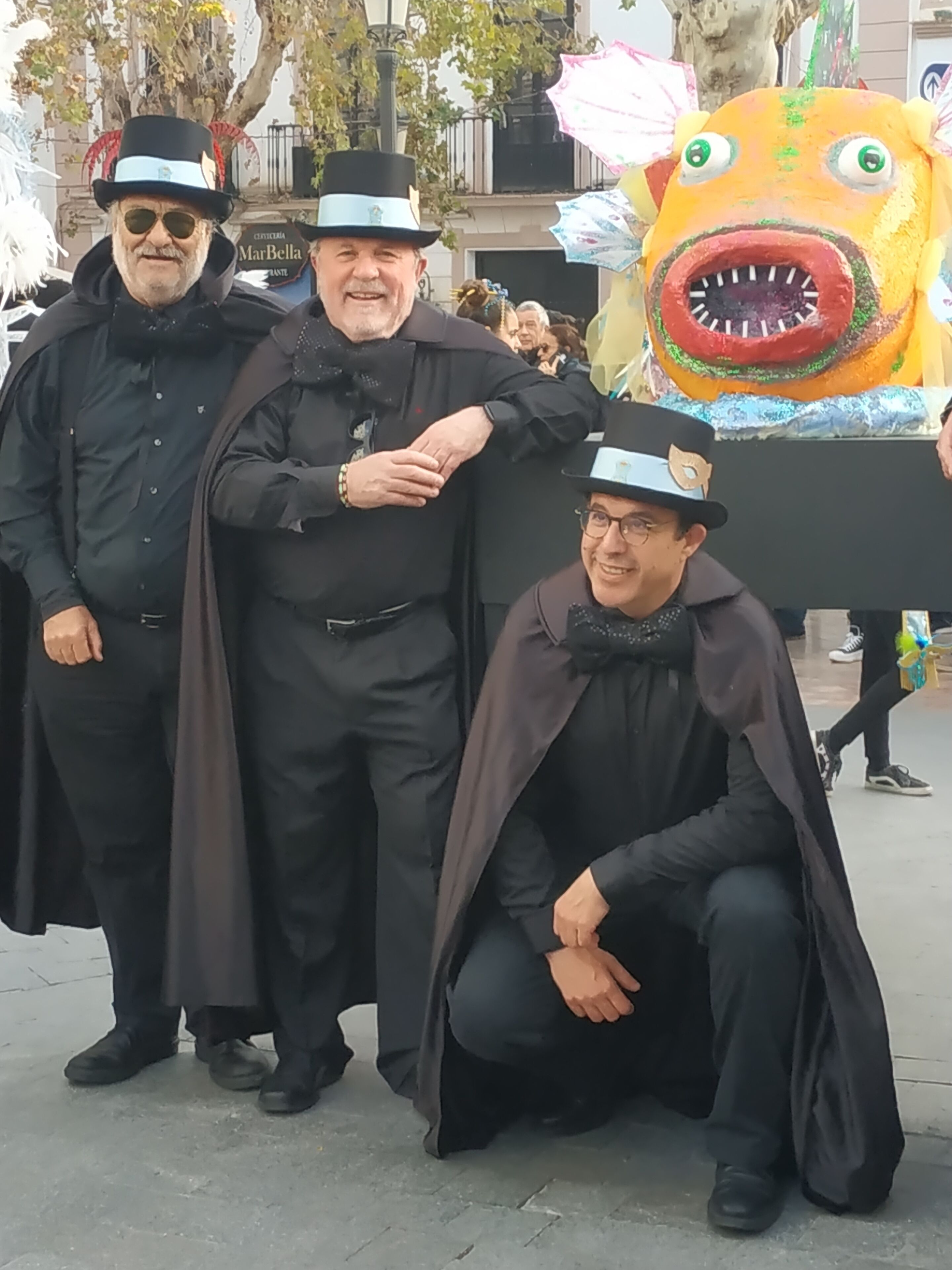 Three individuals in black attire with hats, standing and kneeling in an outdoor setting.