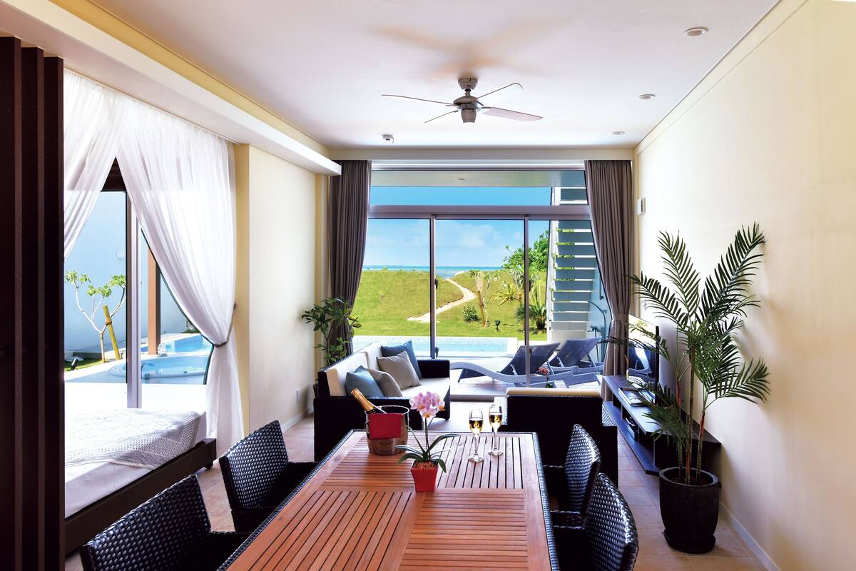 Een kamer met een bed, een eettafel met stoelen, uitzicht op zee en een plafondventilator.