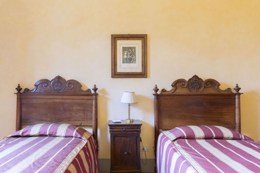 A room with two wooden beds with ornate headboards, a small bedside table with a lamp, and a framed picture on the wall.