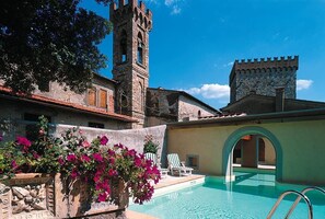 A pool area with a stone wall, a tower, and a building with a tiled roof. - Castle of Volognano (RIGNANO sull arno)