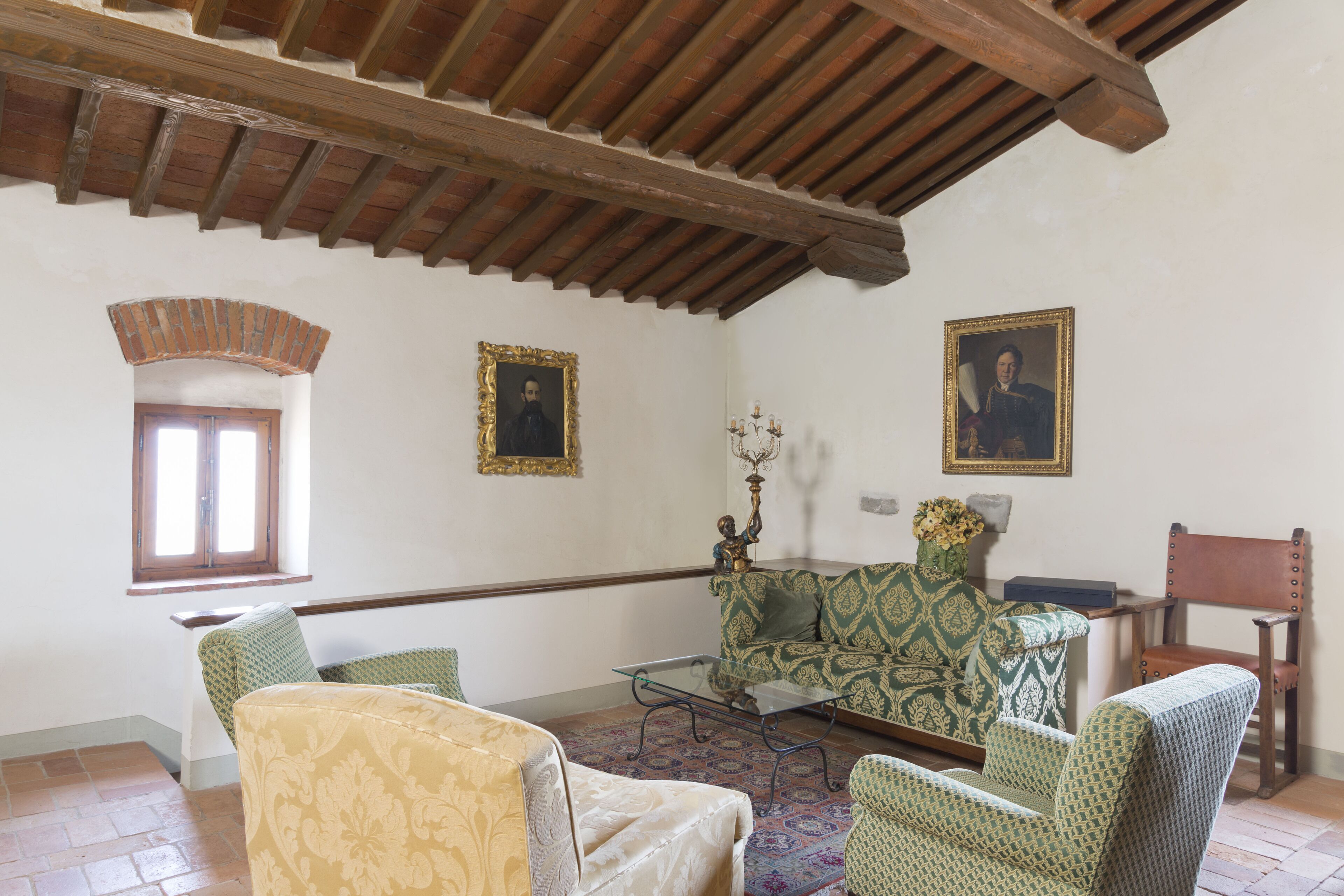 A room with a high wooden ceiling, a patterned sofa, a glass coffee table, and two framed portraits on the wall.