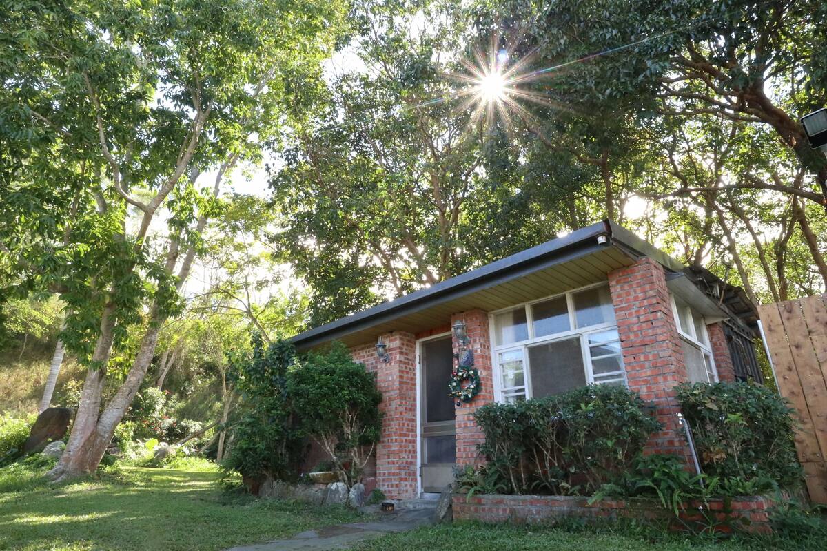 A brick house with a front porch, surrounded by lush greenery and trees.