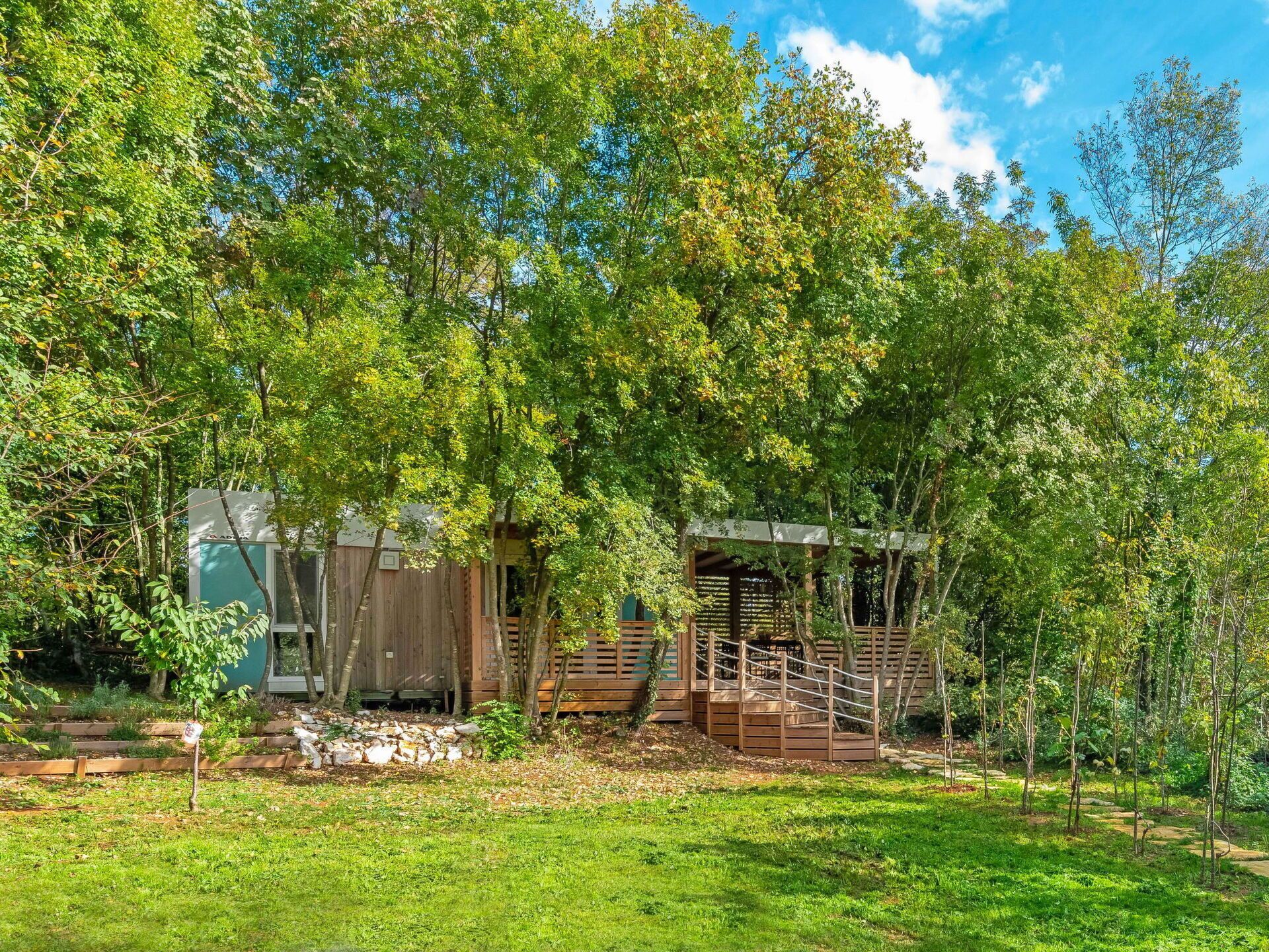 A rustic cabin with a wooden deck and a covered area, surrounded by lush greenery and trees.