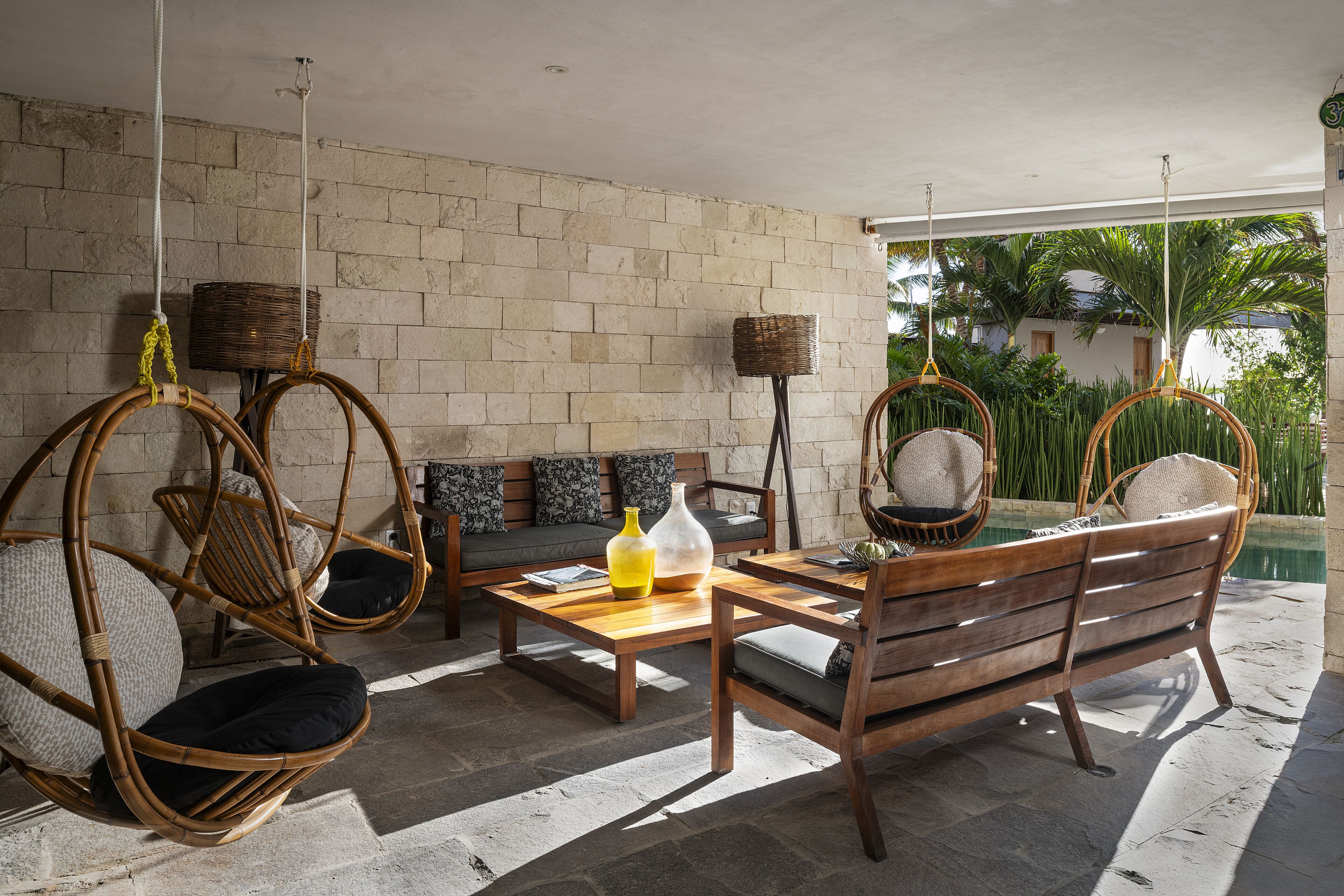 Un espacio exterior acogedor con sillas de mimbre y un banco de madera, una mesa con un jarrón y una lámpara de mimbre.