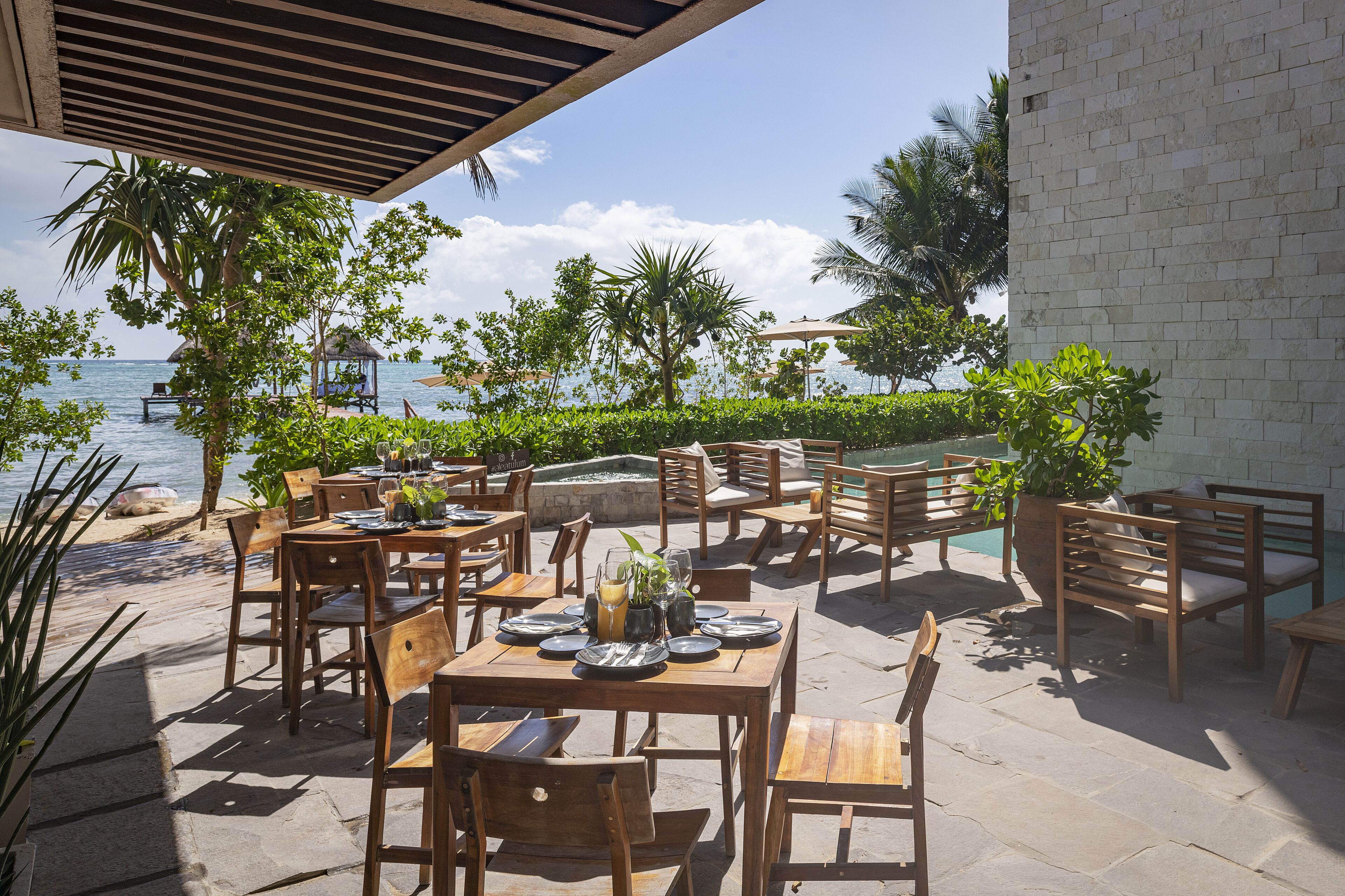 An outdoor dining area with wooden tables and chairs, overlooking a beach and the sea.