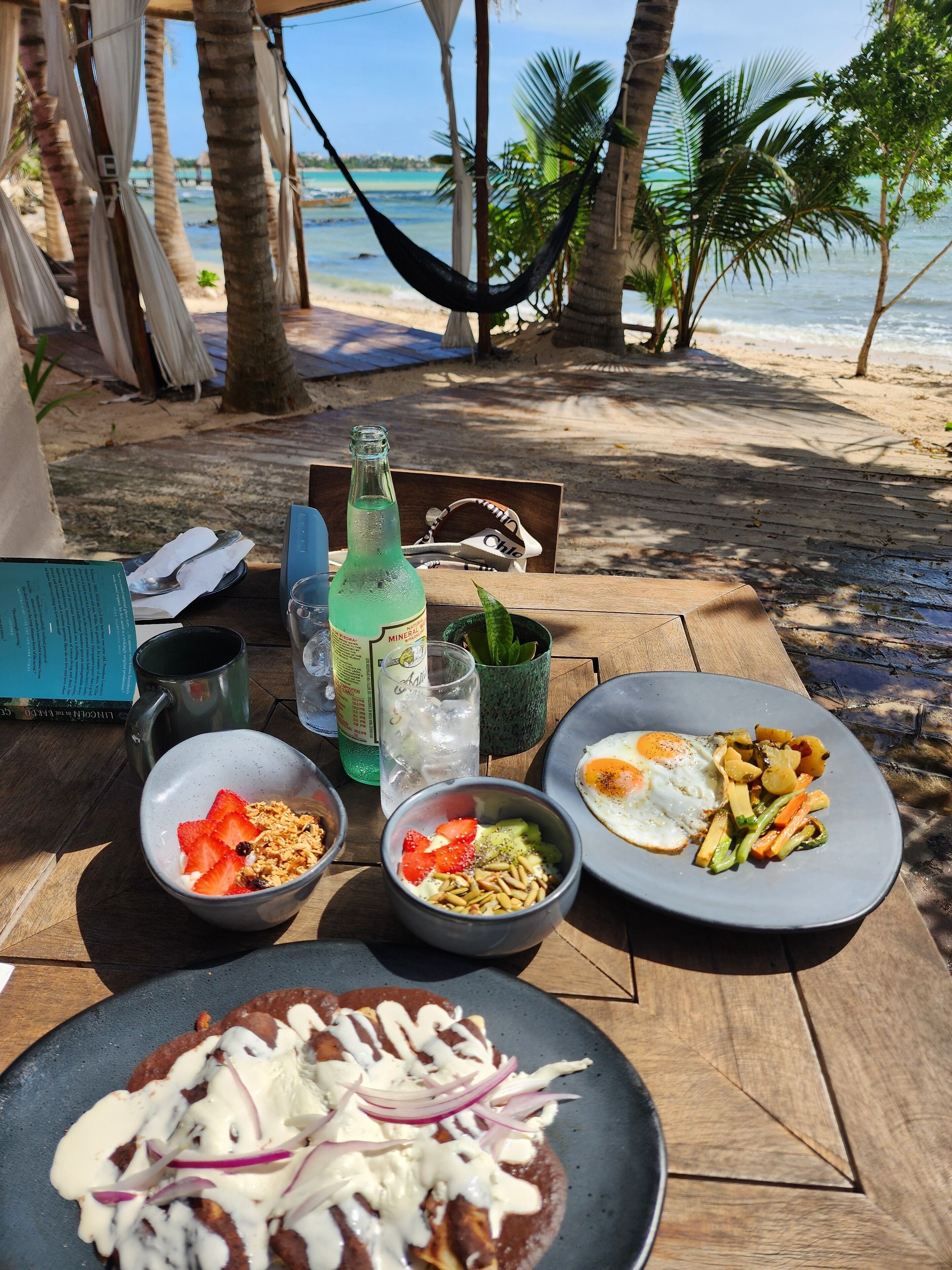 Una mesa de madera con comida que incluye huevos, verduras y una tortilla con ingredientes, sobre una playa con una hamaca y palmeras al fondo.