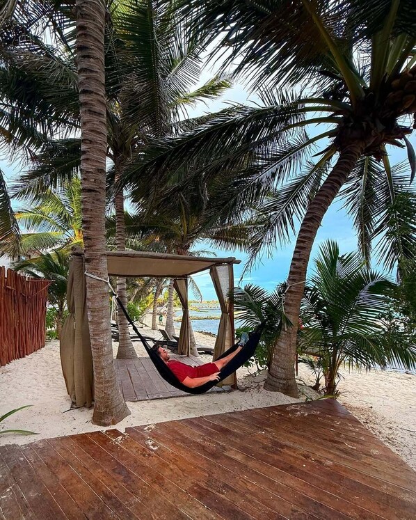 A hammock under a wooden structure on a wooden deck surrounded by palm trees.