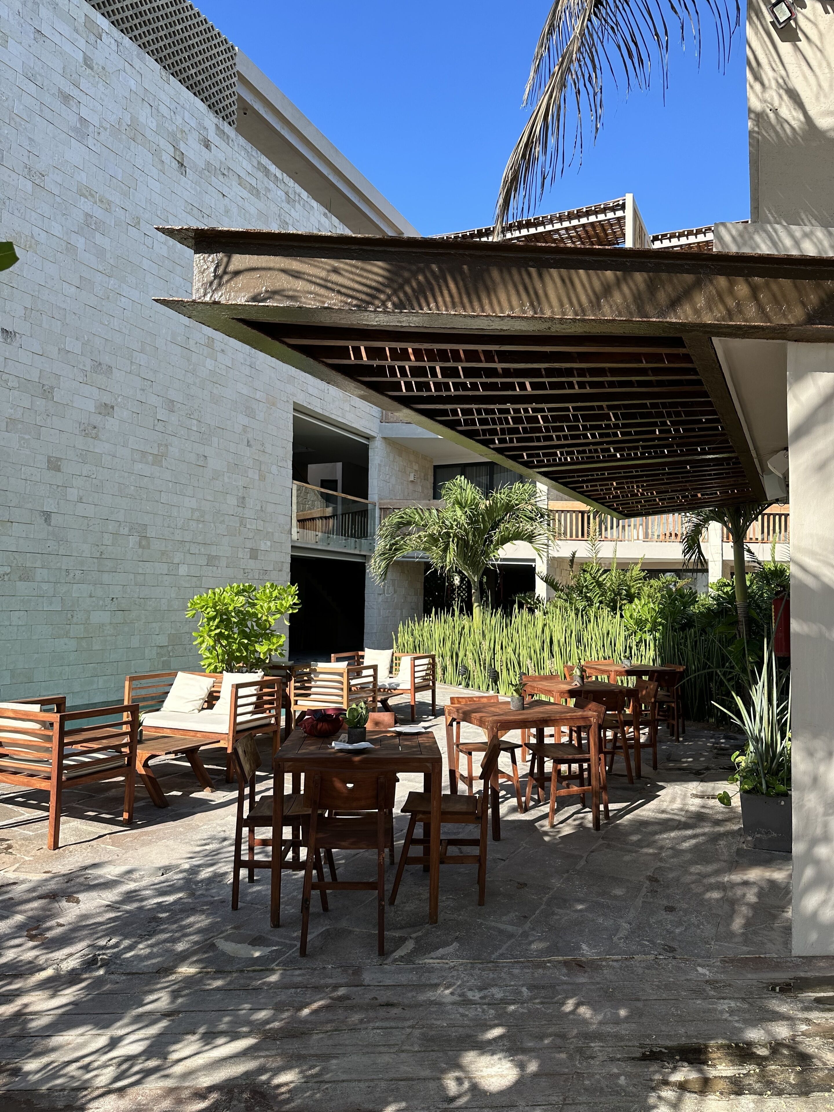 An outdoor patio with wooden furniture and a covered seating area, surrounded by greenery and a white building.
