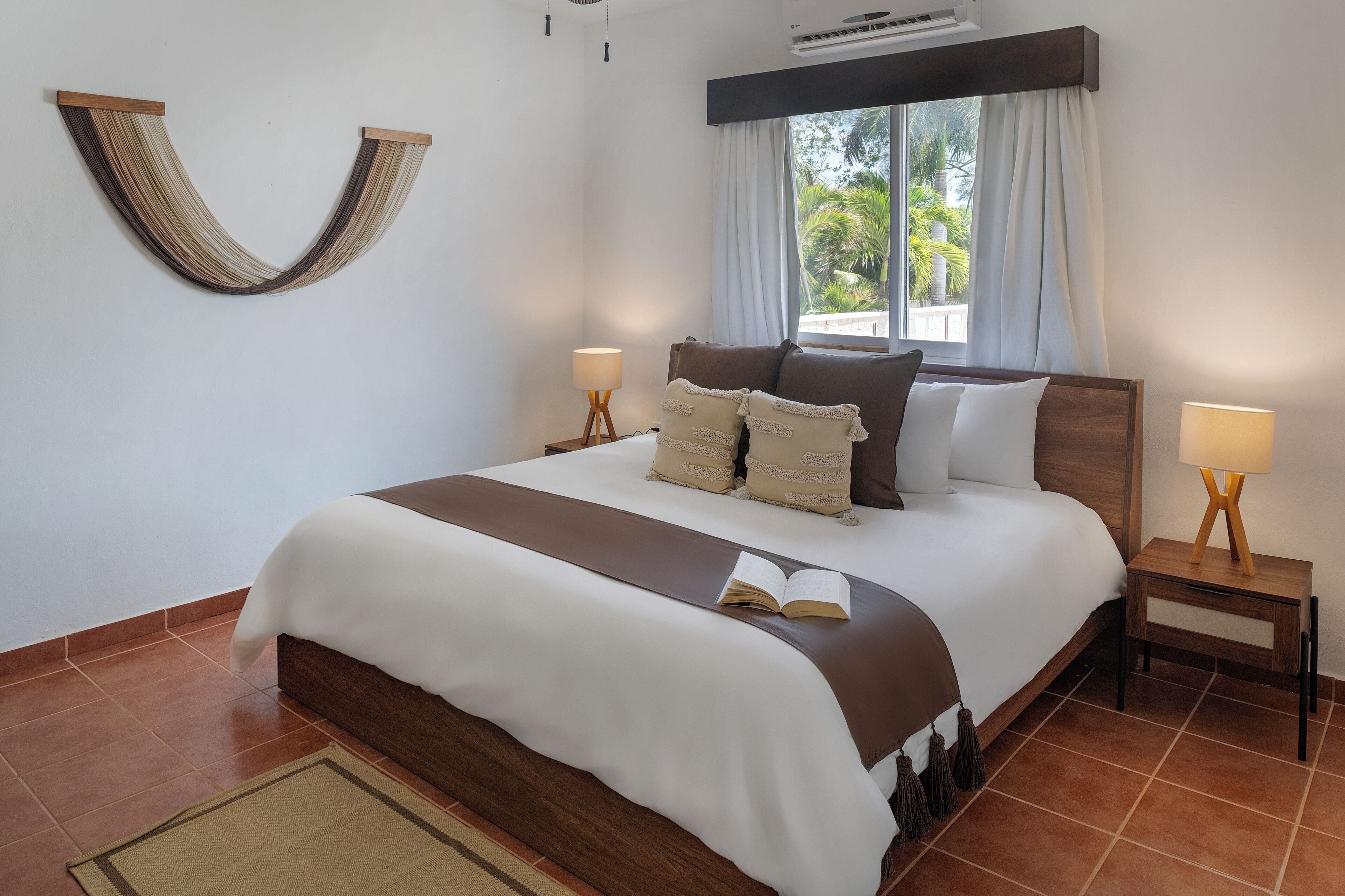 Quarto bem arrumado com uma cama grande, duas mesas de cabeceira com luminárias, uma janela com cortinas e um ponto de luz fixado na parede.