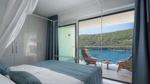 Een slaapkamer met een groot bed, een balkon met rieten stoelen en uitzicht op een meer en heuvels.