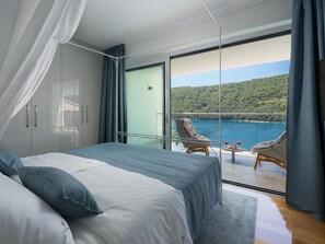 A bedroom with a large bed, a balcony with wicker chairs, and a view of a lake and hills.